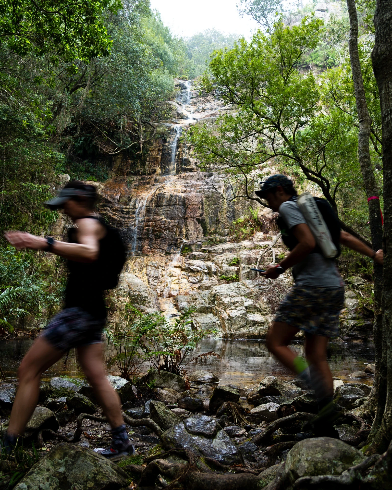 Two trail runners crossing a rocky riverbed with waterfall in the background during the Tsitsikamma race - Photo: Carmen Claire